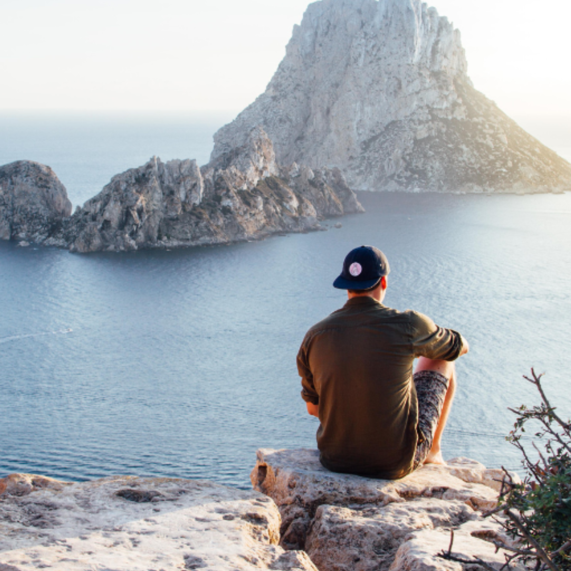 Person sitting on a rocky outcrop overlooking a body of water with a large rock formation.